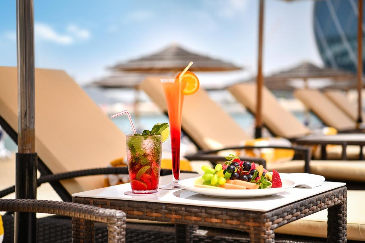 Al Raha Beach Resort & Spa Abu Dhabi Esterno foto The photo shows a table set outdoors, featuring two colorful cocktails and a plate of food. One cocktail is a vibrant red drink garnished with mint, while the other is a layered orange beverage with an orange slice on the rim. The plate of food inclu