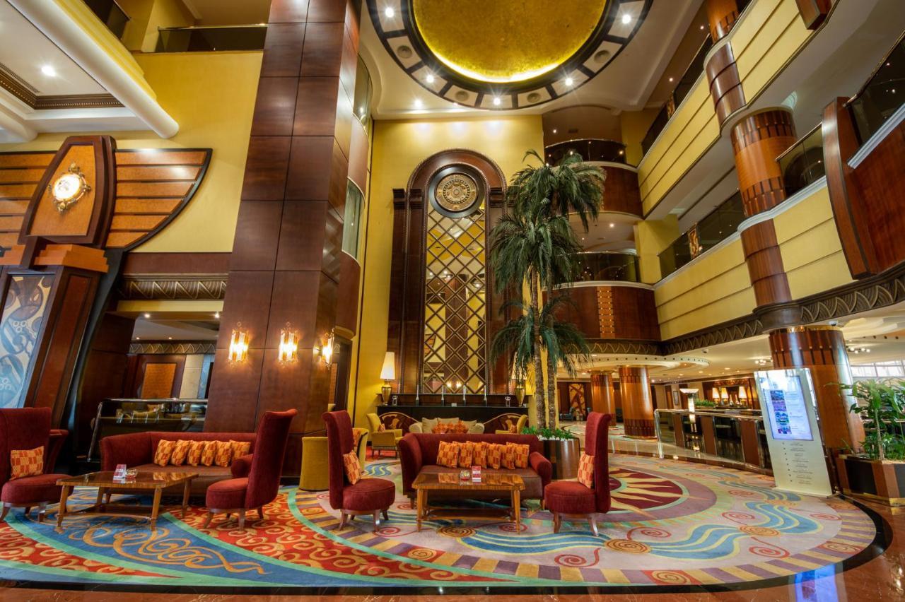 Al Raha Beach Resort & Spa Abu Dhabi Esterno foto The photo shows an elegant lobby interior of a hotel. The space features tall ceilings with decorative lighting and large windows, allowing for natural light. There are several plush seating arrangements, including red armchairs and wooden tables, cr