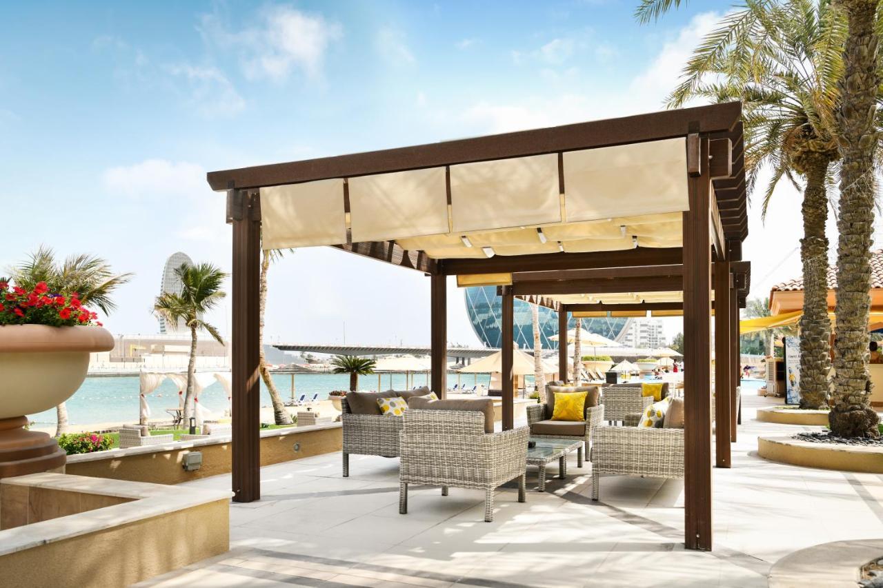 Al Raha Beach Resort & Spa Abu Dhabi Esterno foto The photo shows a stylish outdoor seating area by a beach. There is a wooden pergola covered with a light fabric shade, providing shelter to a set of comfortable wicker chairs and tables. The area is adorned with palm trees, and in the background, th