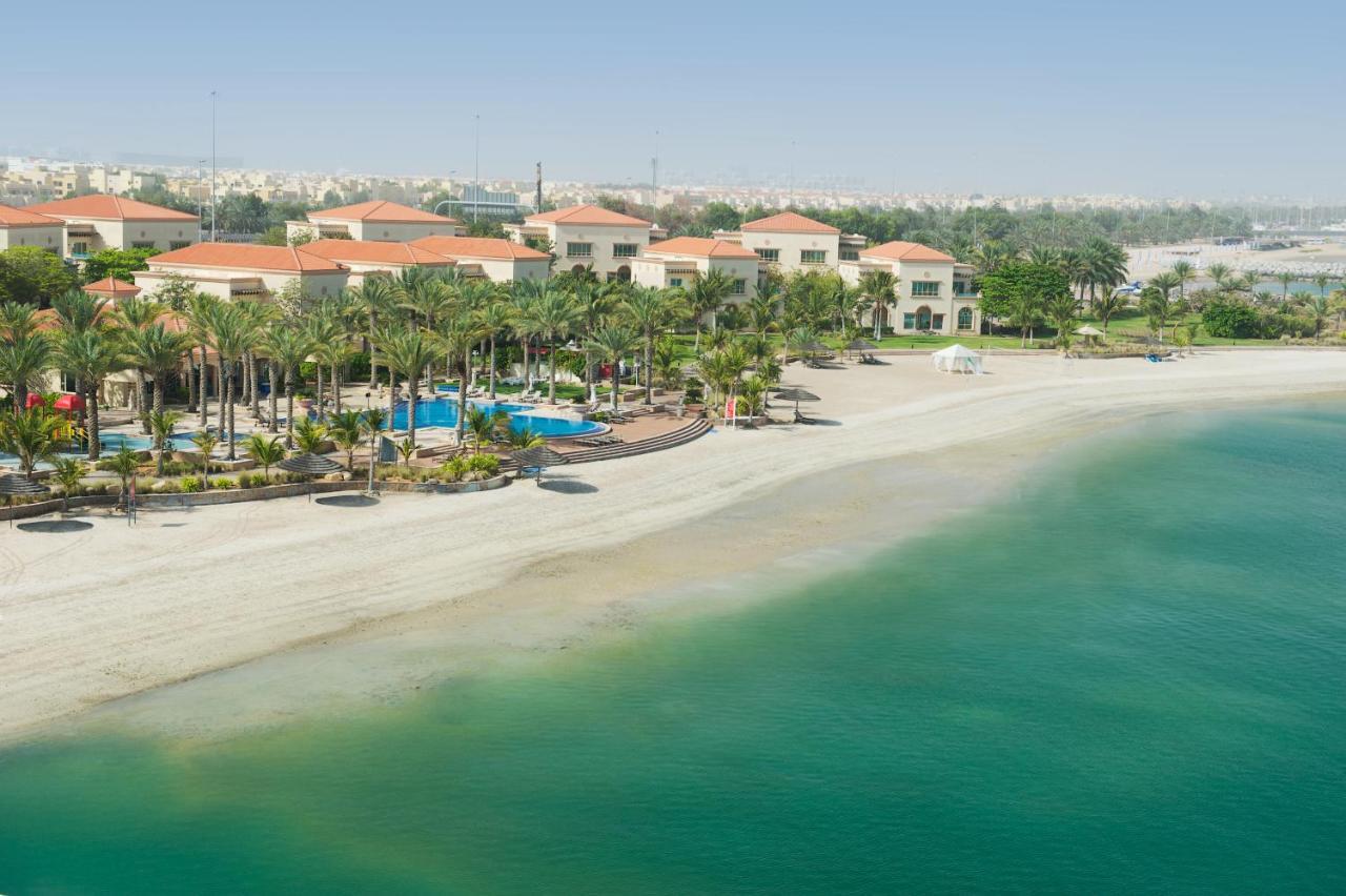 Al Raha Beach Resort & Spa Abu Dhabi Esterno foto The photo depicts a serene beach scene with a clear turquoise sea and a sandy shoreline. In the background, there are several buildings featuring a Mediterranean architectural style with red-tiled roofs. The area is adorned with palm trees and outdoo