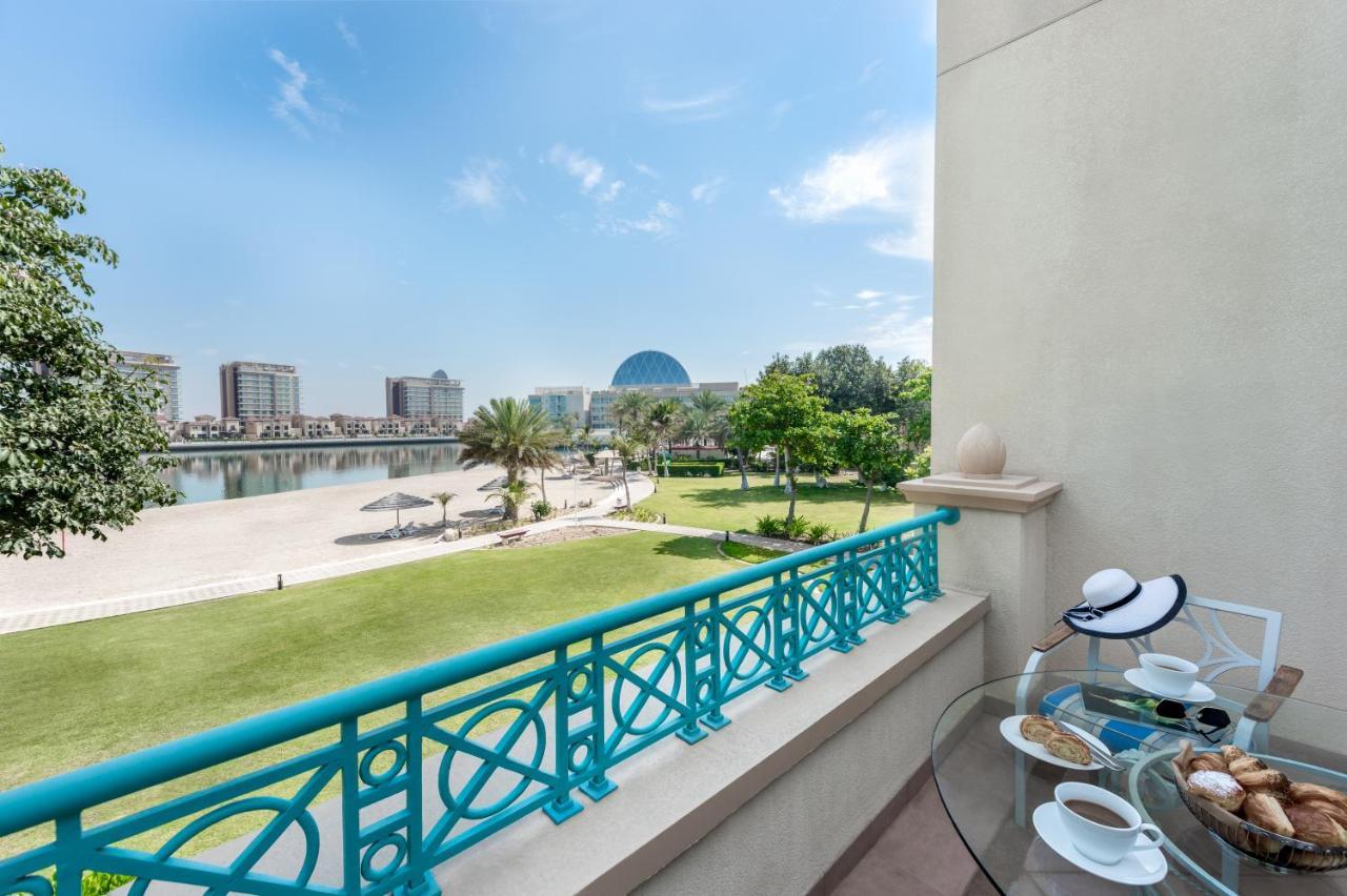 Al Raha Beach Resort & Spa Abu Dhabi Esterno foto The photo shows a balcony view overlooking a scenic landscape. In the foreground, there is a glass table set with snacks or pastries and two cups. Beyond the balcony railing, there is a well-maintained green area with palm trees and a pathway. The ba
