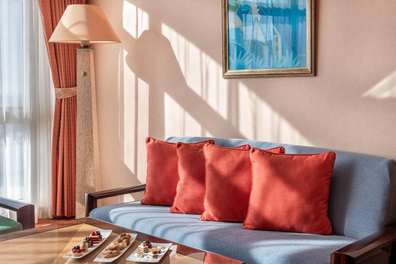Al Raha Beach Resort & Spa Abu Dhabi Esterno foto The photo shows a cozy interior setting, likely a sitting area. It features a light blue couch with four red cushions. In front of the couch, there is a glass table displaying an assortment of snacks or small dishes. The room has soft lighting, with 