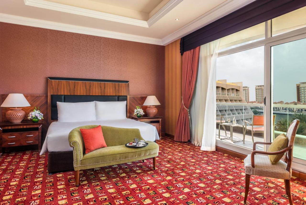 Al Raha Beach Resort & Spa Abu Dhabi Esterno foto The photo shows a hotel room with an elegant and modern design. It features a large bed with white linens and a dark wooden headboard. On either side of the bed are matching bedside lamps. There is a green armchair with a red pillow positioned near t