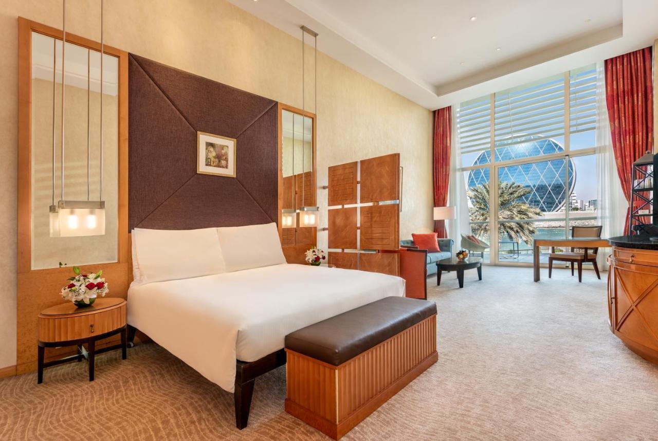 Al Raha Beach Resort & Spa Abu Dhabi Esterno foto The photo shows a modern hotel room featuring a neatly made bed with a white comforter, situated against a stylish headboard with a geometric design. There are bedside tables on either side of the bed, each with a small flower arrangement. A comforta