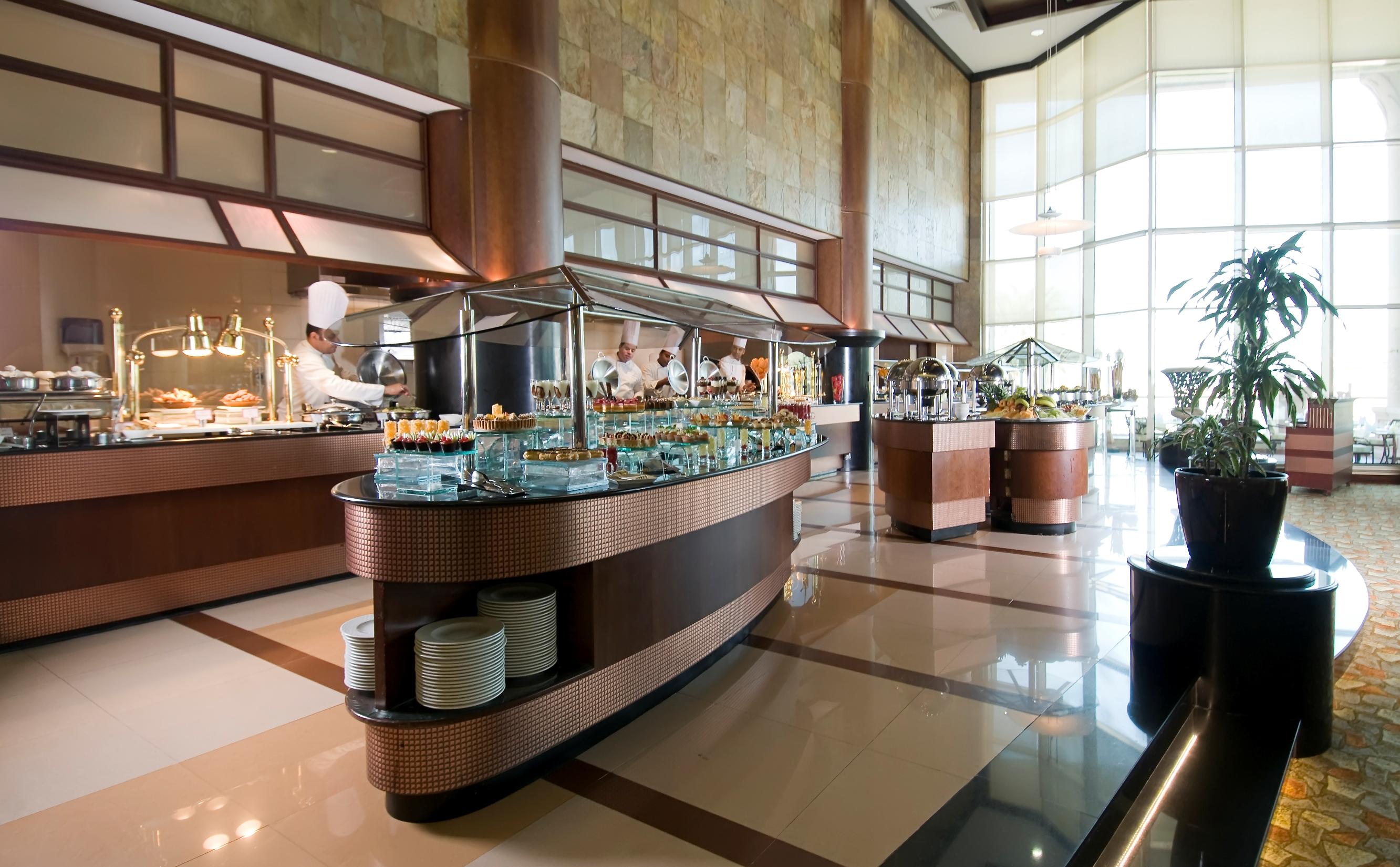 Al Raha Beach Resort & Spa Abu Dhabi Esterno foto The photo shows a spacious buffet area in a restaurant or hotel. There are multiple food stations with glass displays showcasing a variety of dishes. Chefs can be seen in the background, preparing food. The decor features large windows allowing natur