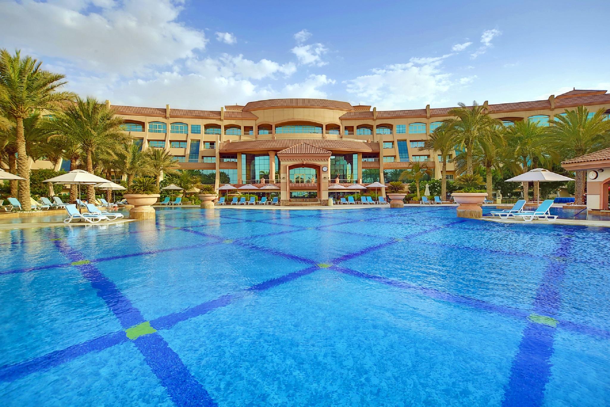 Al Raha Beach Resort & Spa Abu Dhabi Servizi foto The photo features a luxurious resort with a large swimming pool in the foreground. The pool is crystal clear and reflects the sky, bordered by lounge chairs and palm trees. In the background, the resort building showcases modern architecture with la