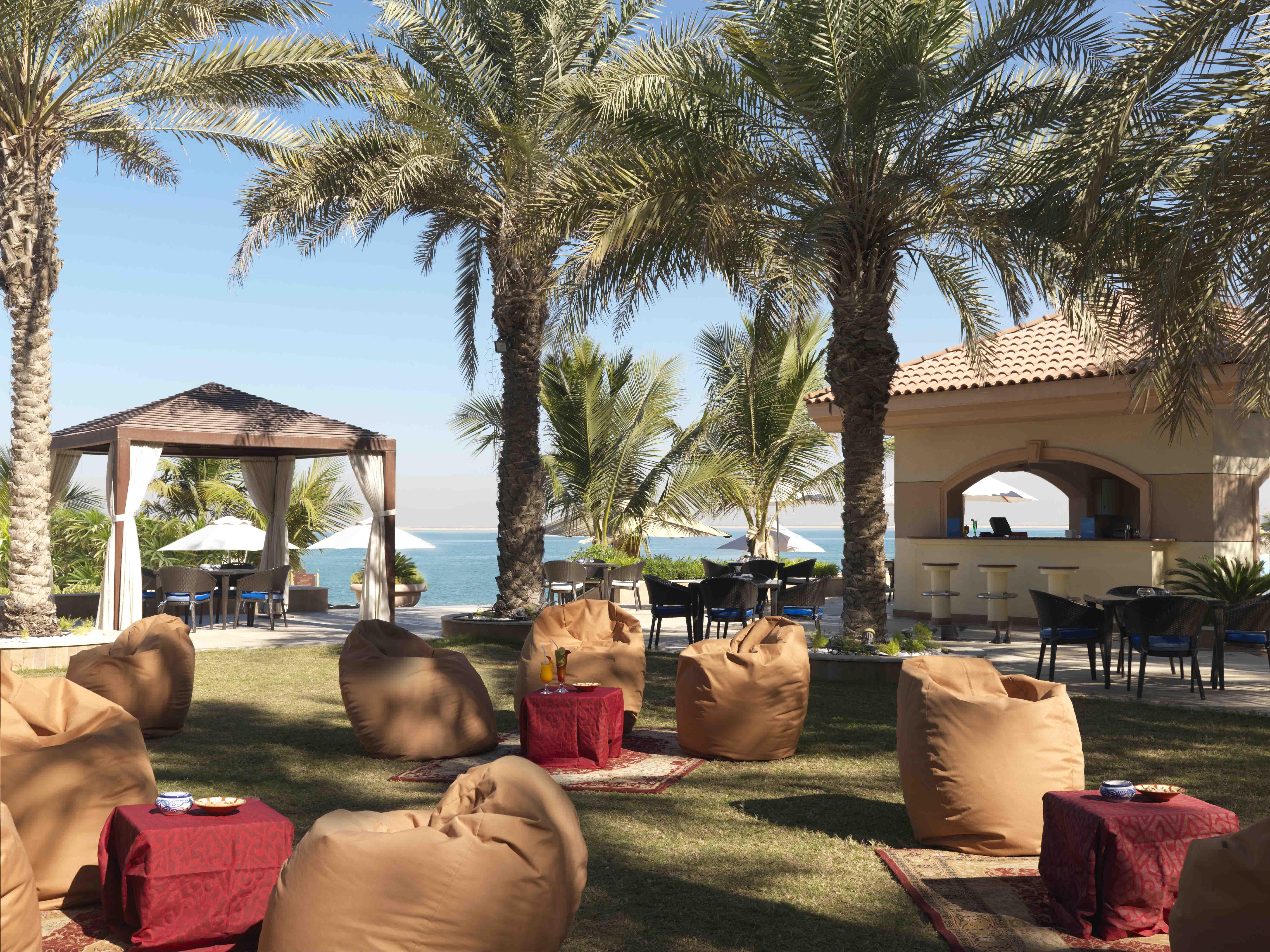 Al Raha Beach Resort & Spa Abu Dhabi Ristorante foto The photo depicts a beautiful outdoor setting with several elements of relaxation and leisure. In the foreground, there are large, comfortable bean bag chairs arranged on a grassy area, accompanied by low tables. Surrounding this seating area are tal