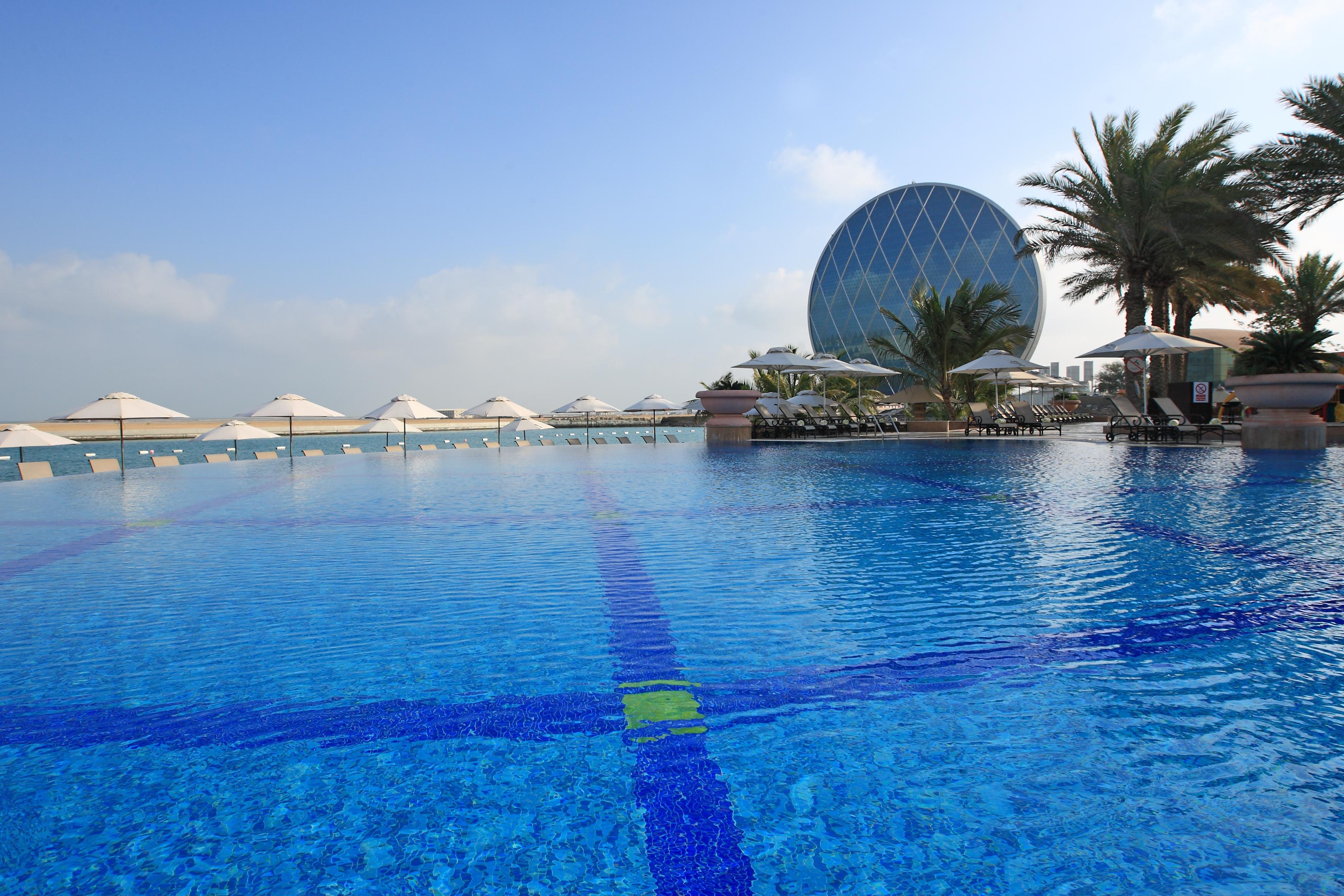 Al Raha Beach Resort & Spa Abu Dhabi Esterno foto The photo features a large, serene swimming pool with clear, blue water reflecting the sky. Surrounding the pool are several lounge chairs and umbrellas, suggesting a relaxed, resort-like atmosphere. In the background, a distinctive building with a r
