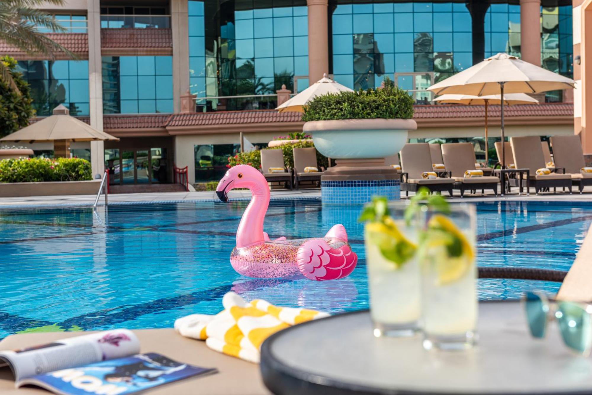 Al Raha Beach Resort & Spa Abu Dhabi Esterno foto The photo depicts a bright and inviting outdoor swimming pool surrounded by a luxurious setting. In the pool, there is a large pink flamingo float, adding a fun and whimsical touch. To the side, a table is set up with two refreshing drinks, likely co