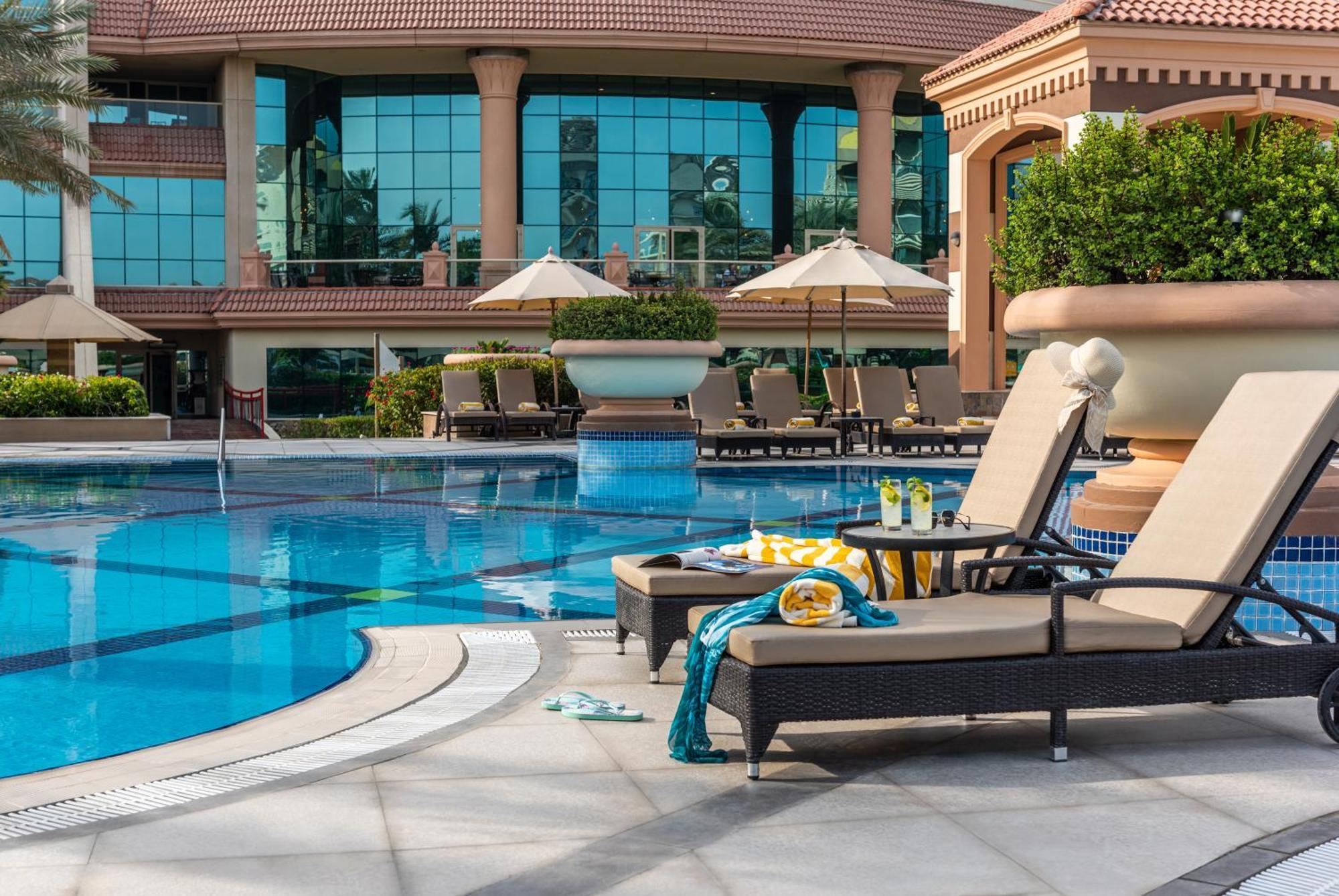 Al Raha Beach Resort & Spa Abu Dhabi Esterno foto The photo depicts a luxurious outdoor swimming pool area. In the foreground, there are two lounge chairs with cushions, positioned beside the pool. A small table between them holds drinks and snacks, suggesting a relaxing atmosphere. The pool itself 