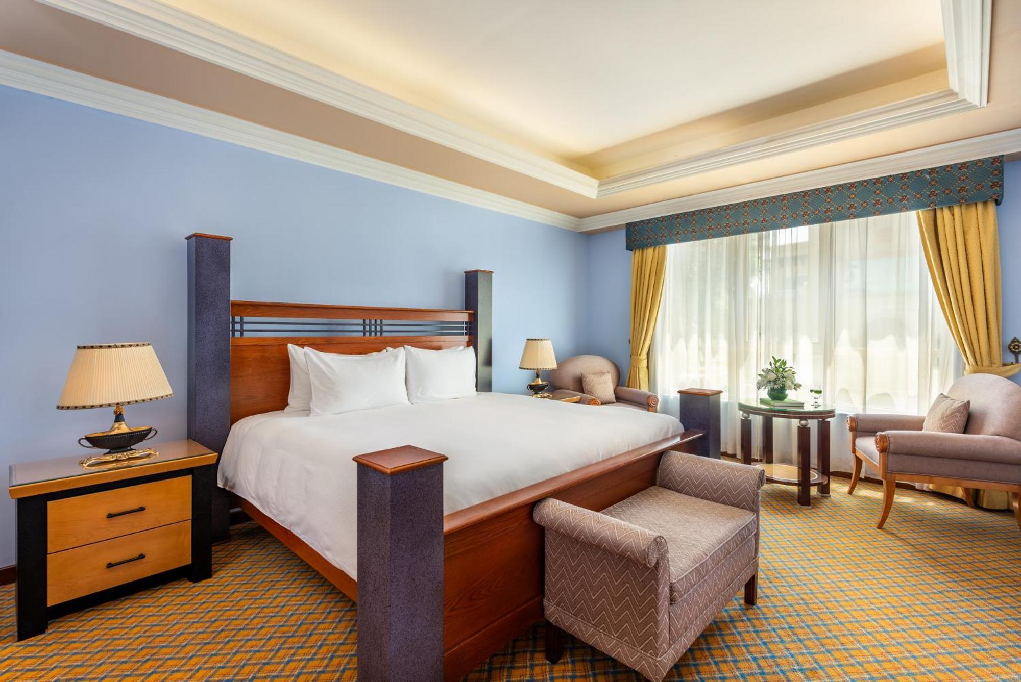 Al Raha Beach Resort & Spa Abu Dhabi Esterno foto The photo shows a well-decorated hotel room. It features a large bed with a wooden frame, neatly made with white bedding. On either side of the bed are bedside tables with lamps. There is a comfortable chair next to a window that has sheer curtains a
