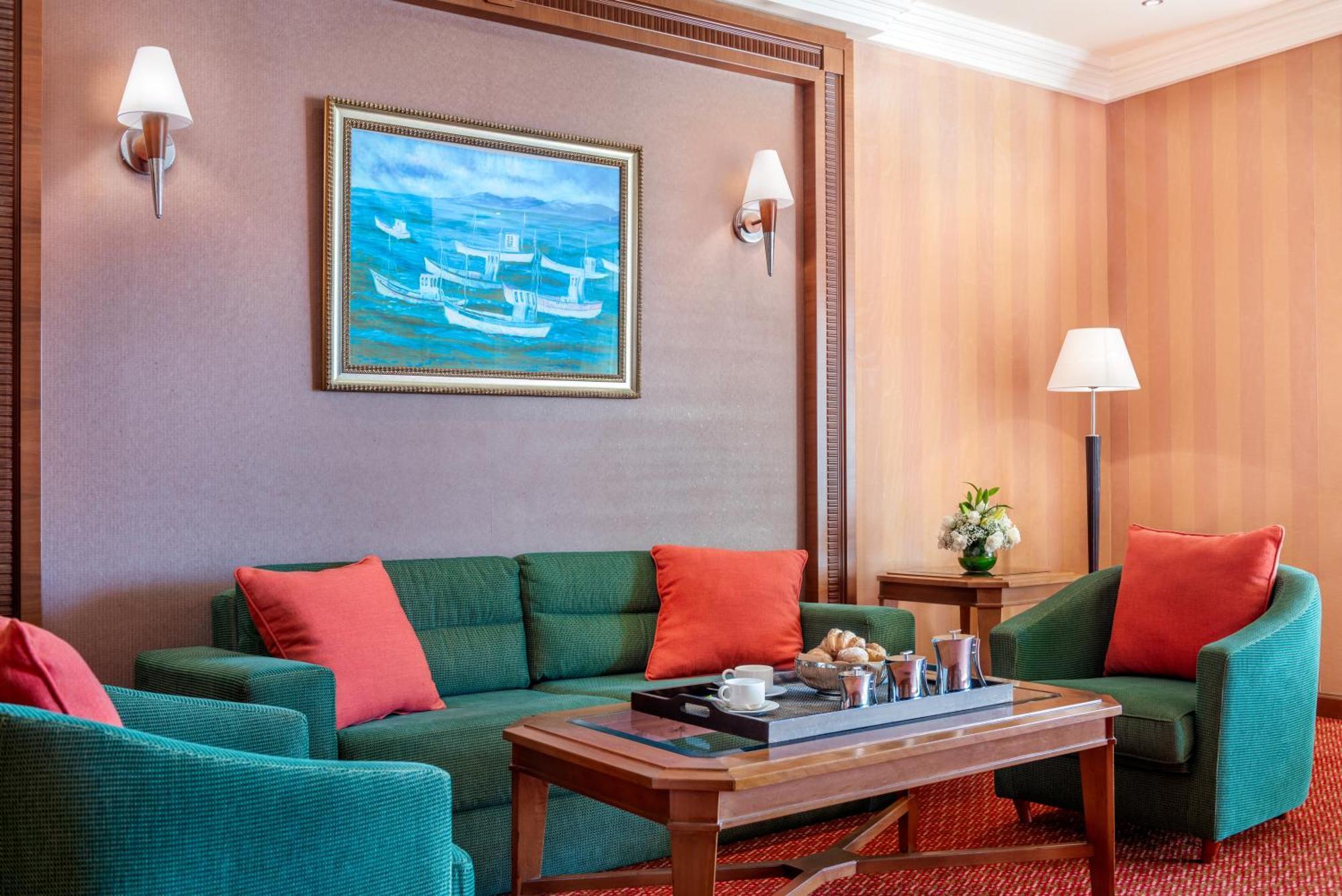Al Raha Beach Resort & Spa Abu Dhabi Esterno foto The photo displays a cozy and elegantly furnished living area. It features a green sofa adorned with orange cushions and two matching green armchairs. A wooden coffee table sits in the center, which holds a tray with beverages and snacks. The walls a