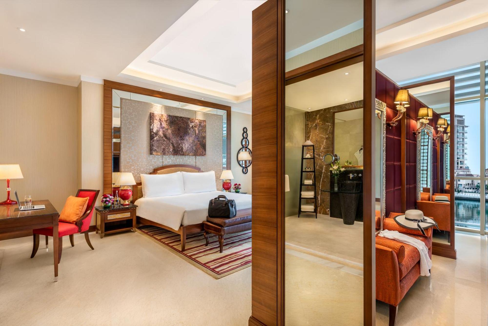 Al Raha Beach Resort & Spa Abu Dhabi Esterno foto The photo shows a luxurious hotel room with a modern design. The room features a large bed with white linens, flanked by bedside tables with lamps. There is a area with a chair and a small table on the left side, and a vibrant artwork hanging above t