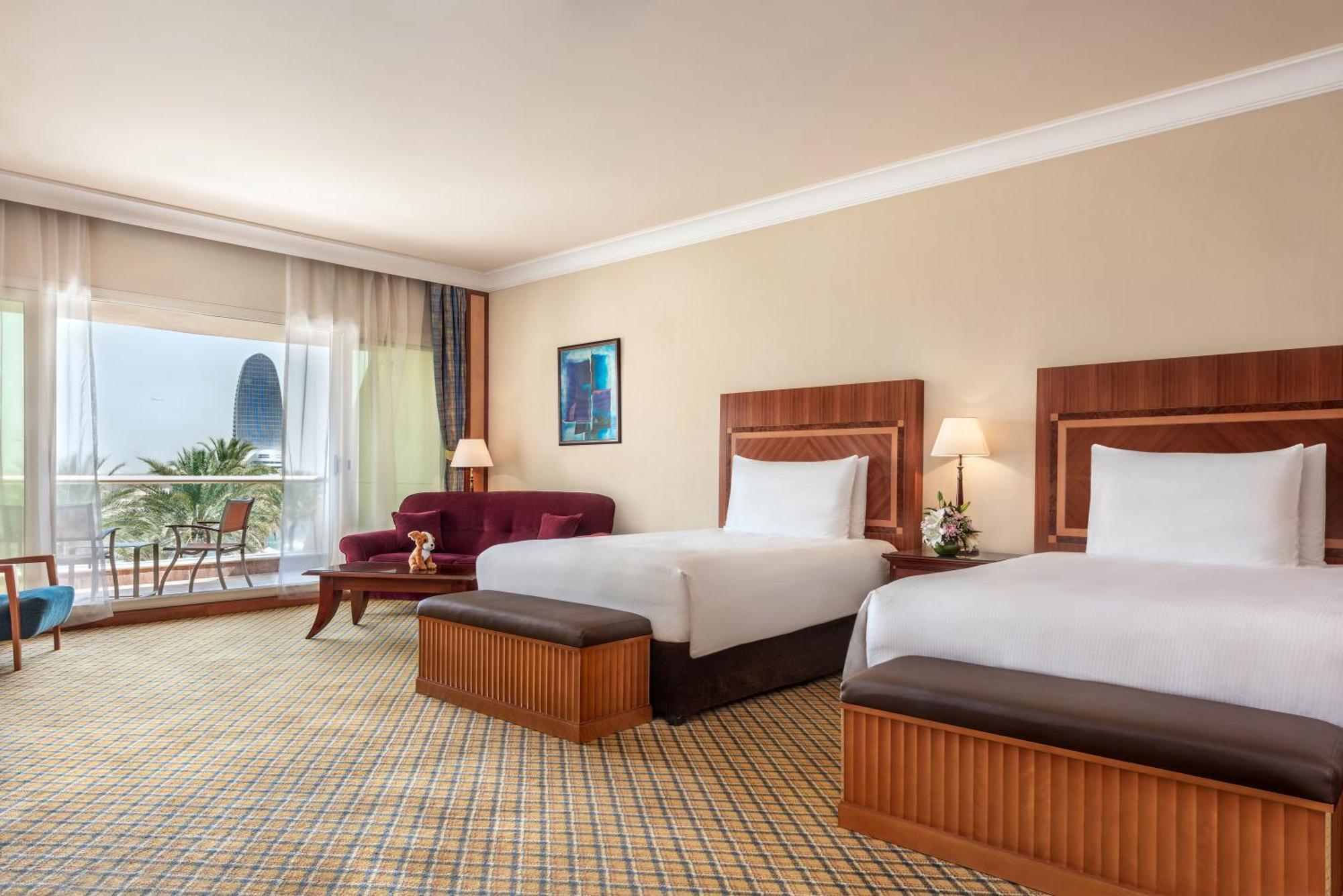 Al Raha Beach Resort & Spa Abu Dhabi Camera foto The photo shows a hotel room featuring two twin beds with wooden headboards. The room has a light, neutral color scheme, with a large window that provides natural light and a view of the outdoors. There is a small seating area with a purple sofa and 