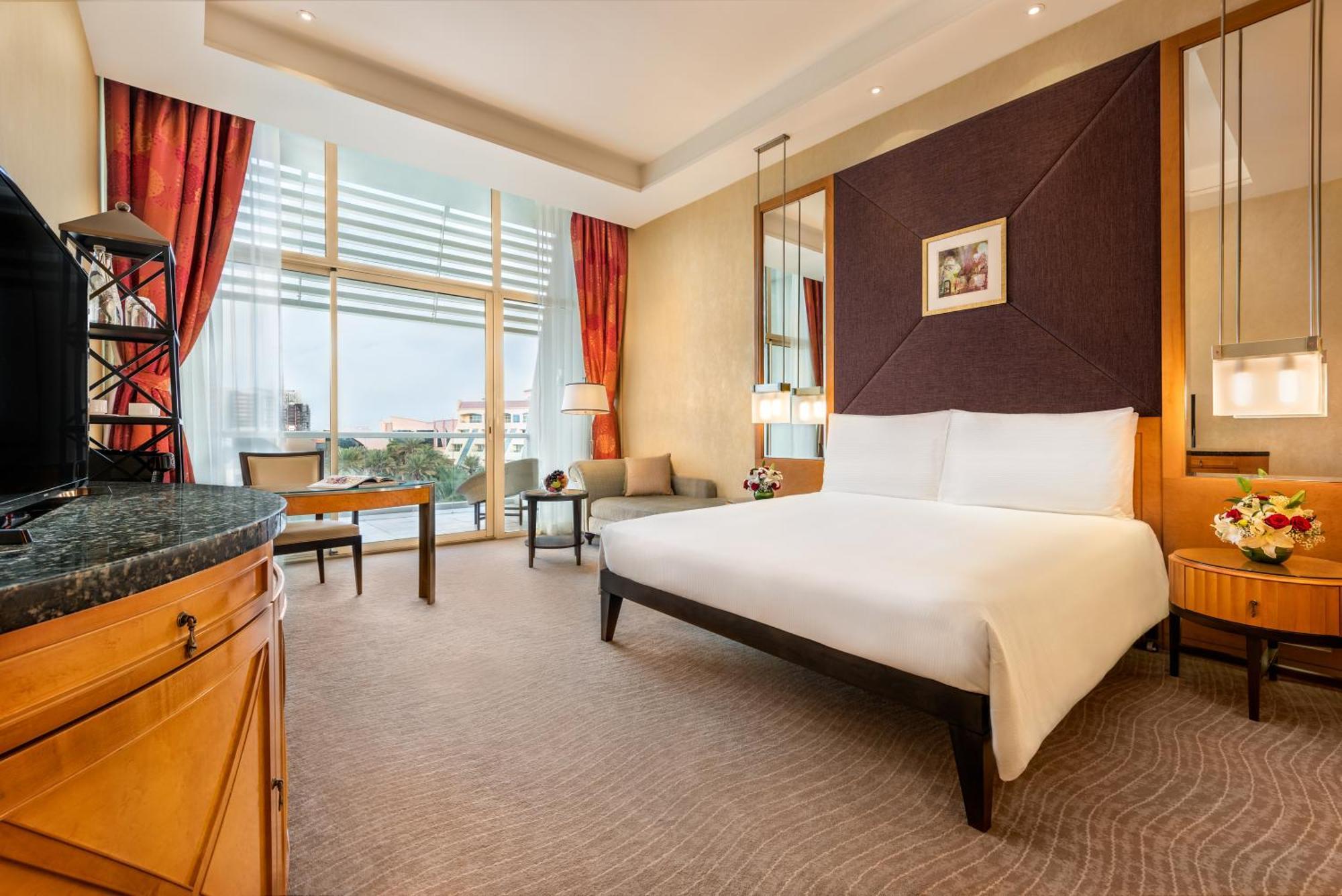 Al Raha Beach Resort & Spa Abu Dhabi Camera foto The photo shows a modern hotel room featuring a large bed with a white duvet positioned against a purple accent wall. There are tall windows with sheer curtains allowing natural light to enter the space, along with red drapes framing the windows. A d