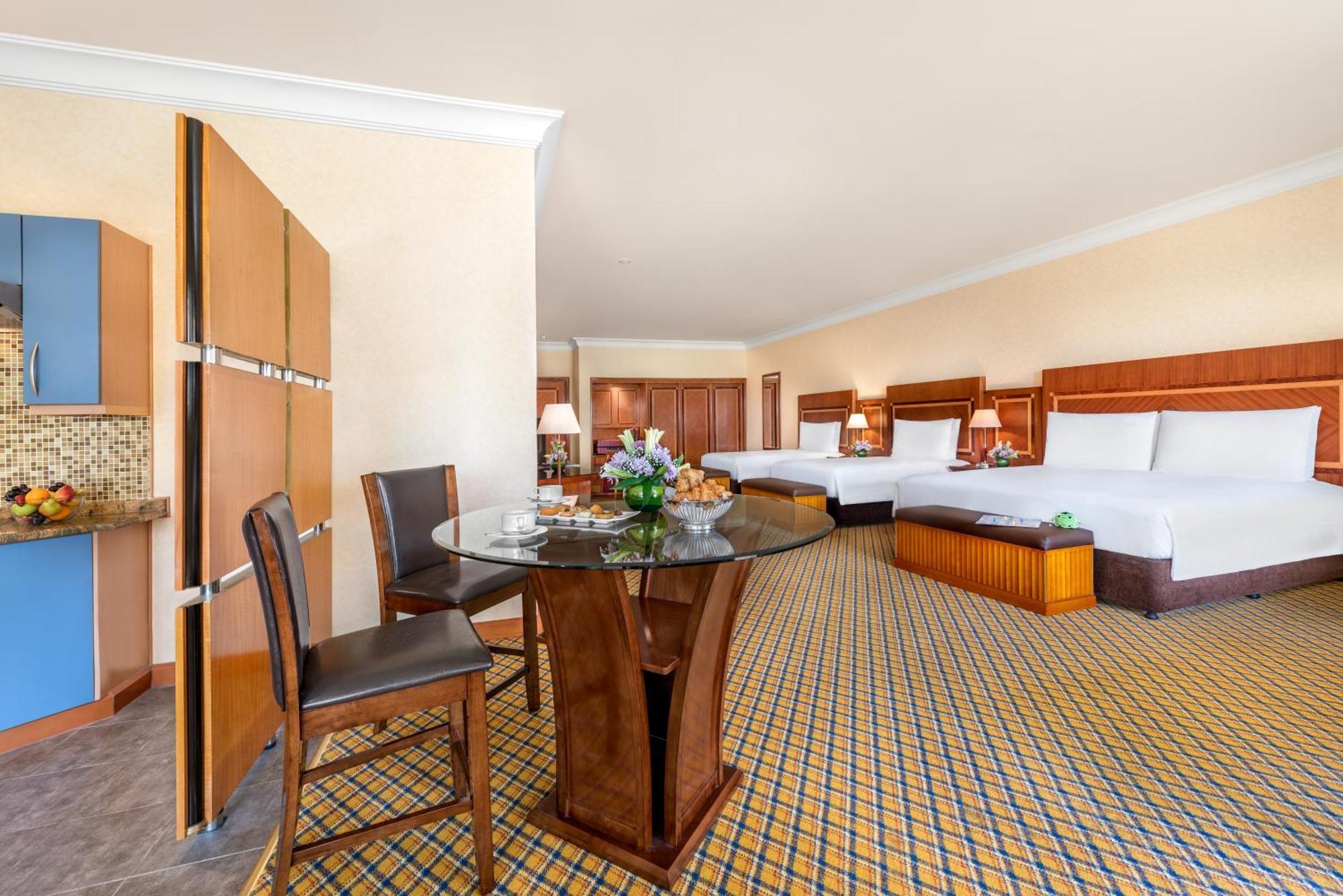 Al Raha Beach Resort & Spa Abu Dhabi Camera foto The photo displays a spacious hotel room. It features two large beds with white bedding, a circular glass dining table with a decorative floral centerpiece, and a couple of chairs around it. There is a wooden cabinet along one wall, and the room has 
