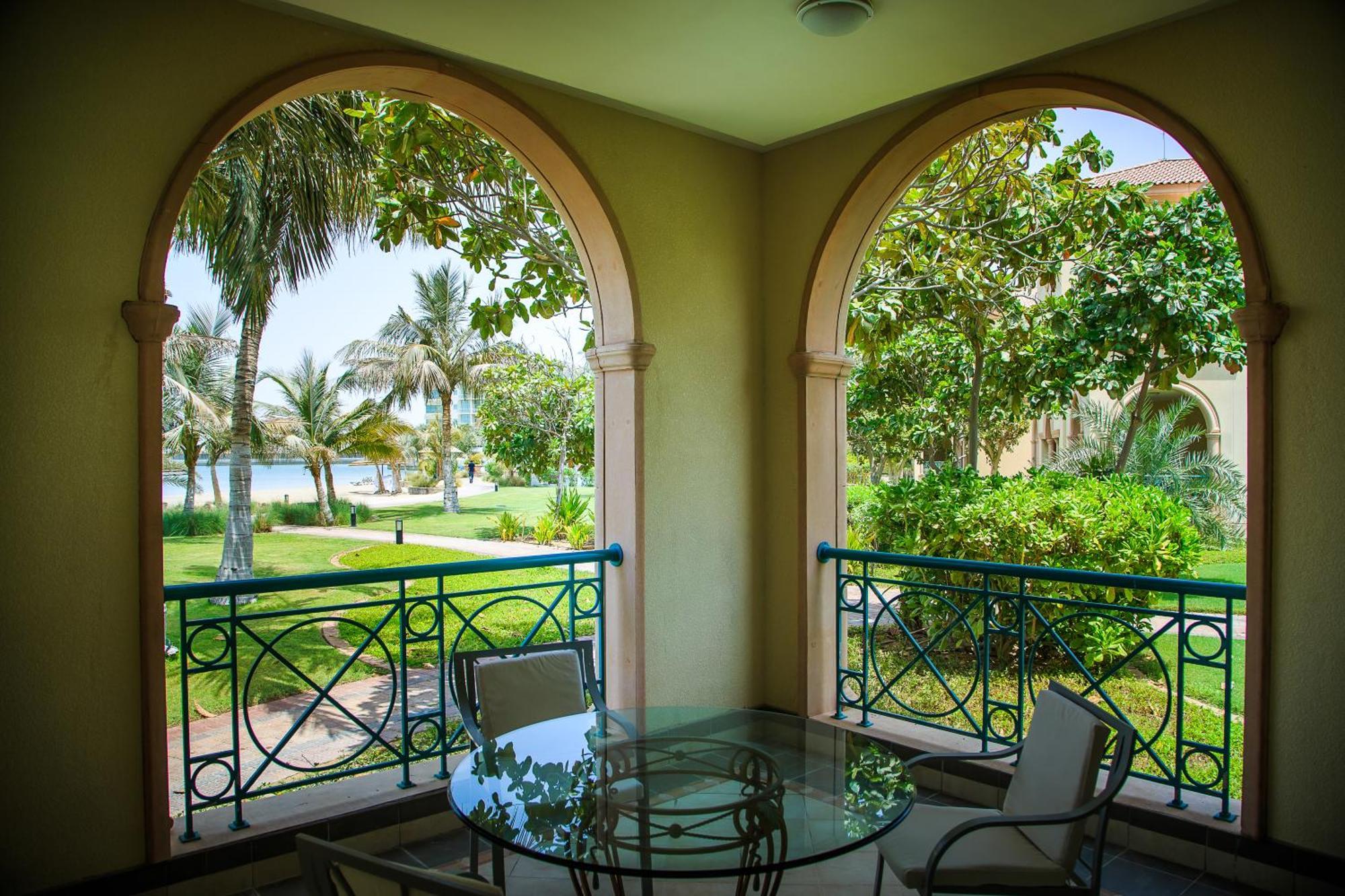 Al Raha Beach Resort & Spa Abu Dhabi Camera foto The photo displays a beautifully designed balcony or terrace area. It features two large arches that frame the view outside. Through the arches, you can see a lush garden with palm trees and well-maintained greenery. There’s a glass table with severa