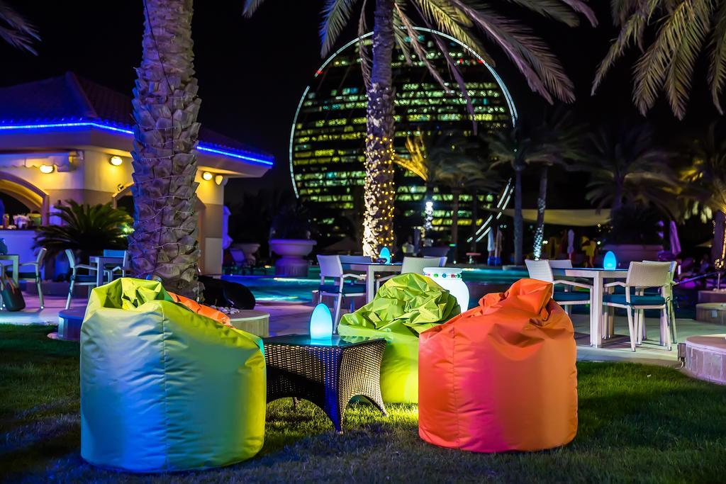 Al Raha Beach Resort & Spa Abu Dhabi Esterno foto The photo shows a vibrant outdoor lounge area at night. In the foreground, there are colorful bean bag chairs—one in bright green, one in yellow, and one in orange. A small table sits between the bean bags. In the background, illuminated palm trees a