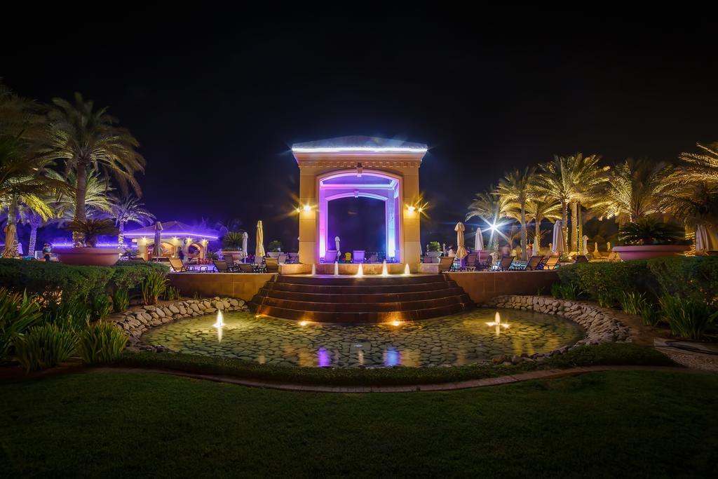 Al Raha Beach Resort & Spa Abu Dhabi Esterno foto The photo depicts a beautifully lit outdoor scene at night. In the foreground, there is a serene water feature with a circular pond, and small fountains creating gentle ripples. Surrounding the pond are lush green lawns and ornamental plants. 

In th