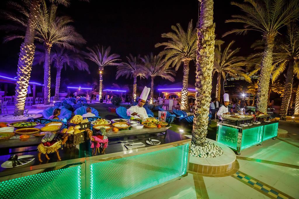 Al Raha Beach Resort & Spa Abu Dhabi Esterno foto The photo depicts an outdoor dining area at night, featuring several chefs in white uniforms and hats working at a food station. The setting is illuminated by colorful lights, with glowing green accents on the serving tables. Surrounding the area are